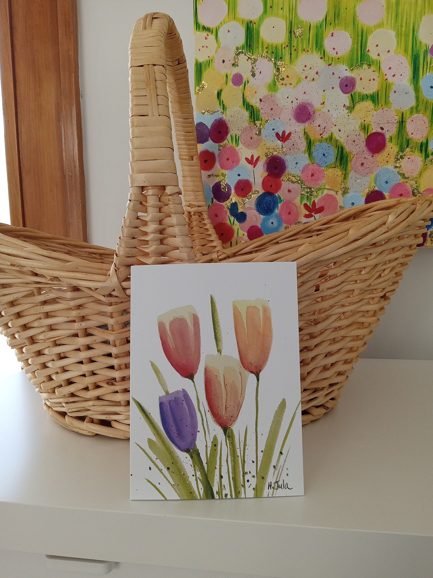 Wicker basket with a floral painting and a card with tulips on a white surface