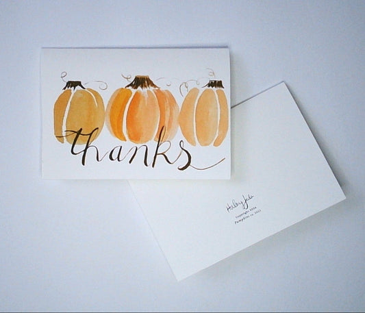A white greeting card with a watercolor illustration of three orange pumpkins and the word 'thanks' in brown script, shown with a backside view which displays Artist's signature